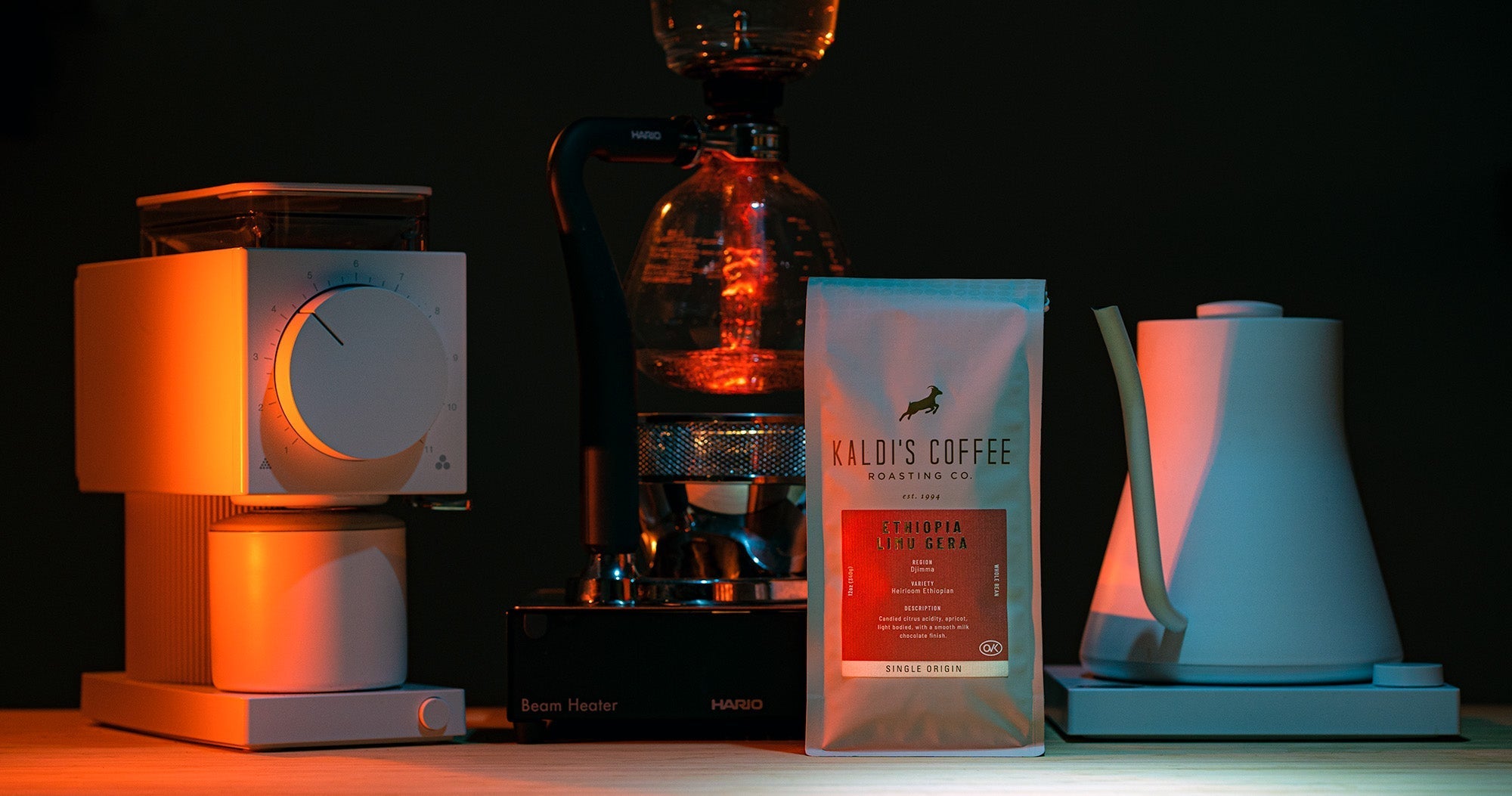 Coffee brewing set up featuring a grinder, a siphon coffee maker, a bag of coffee, and a kettle