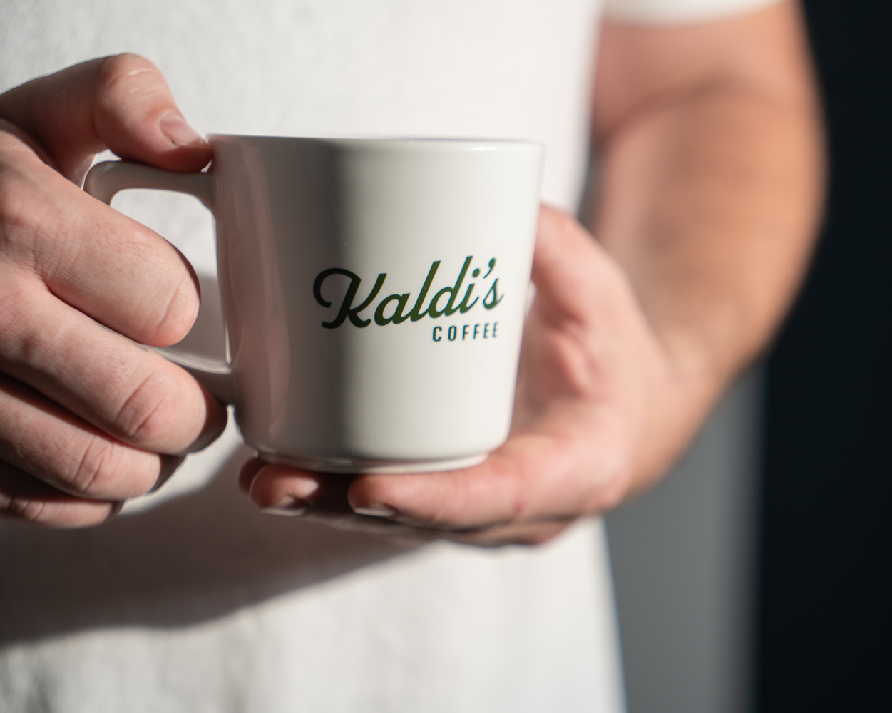 A person holding a white ceramic coffee mug with 'Kaldi's Coffee' printed on it.