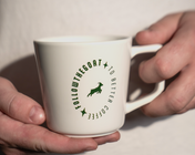 A white ceramic coffee mug with a green circular graphic reading 'FOLLOW THE GOAT TO BETTER COFFEE'.