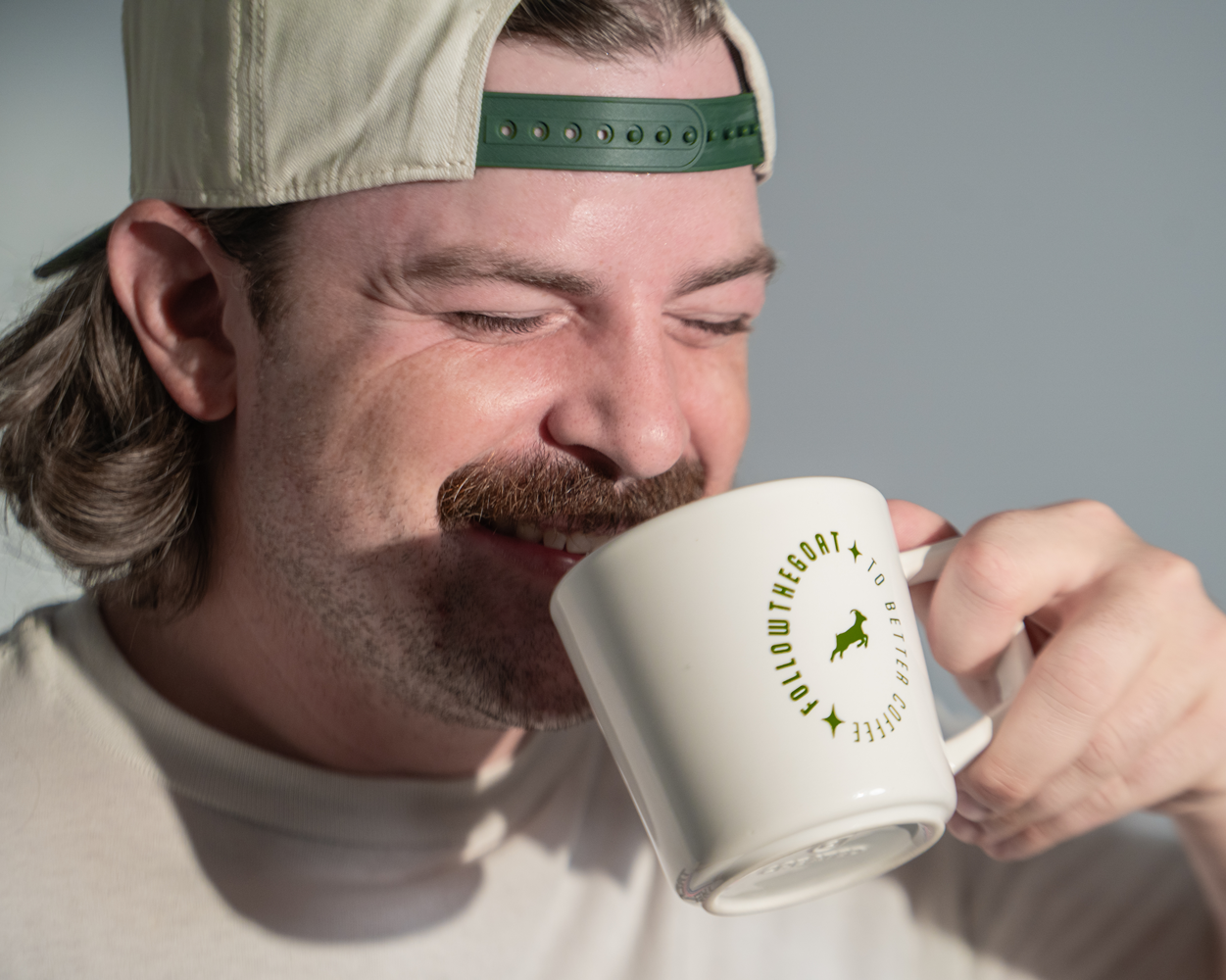 A man drinks from a white ceramic mug featuring a green circular logo with a goat silhouette and text.