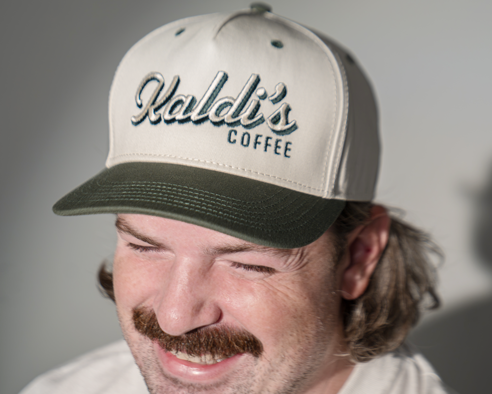 A close-up of a man wearing a white and dark green baseball cap with 'Kaldi's Coffee' embroidered on the front.