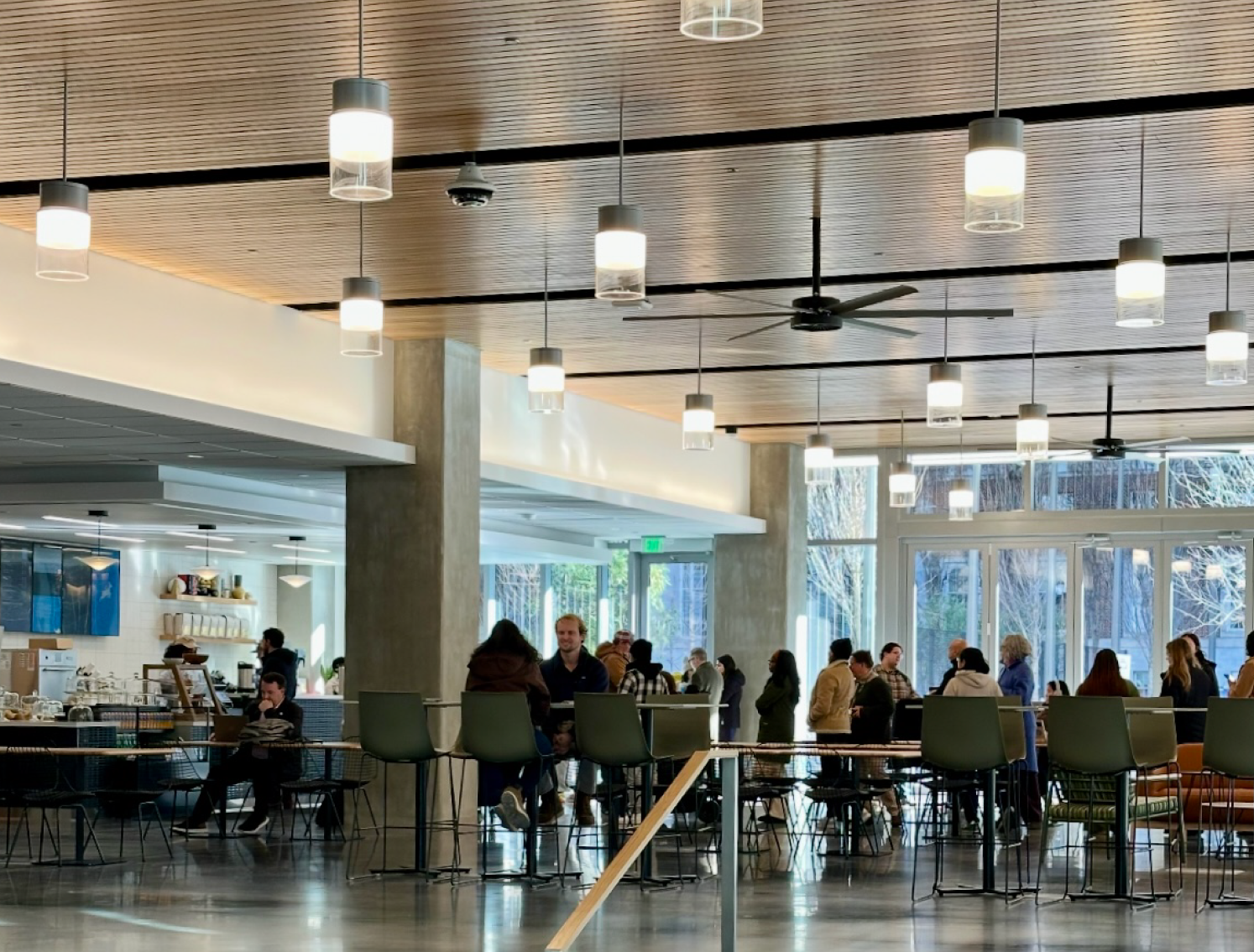 Modern office cafeteria with people sitting at tables, large windows, and modern decor.