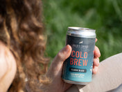 Close-up of a hand holding a chilled can of Kaldi's Coffee Cold Brew, Classic Black. The can features pink "COLD BREW" lettering.